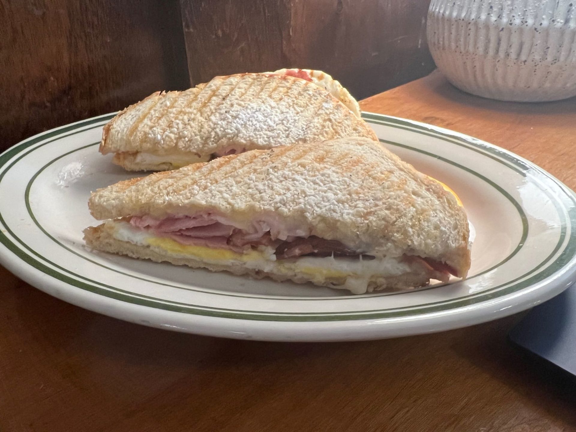 Two halves of a sandwich, cut diagonally, rest on an oval shaped white plate. There is ham, cheese, and egg melted together in the center, and the tops of the bread are dusted with powdered sugar.
