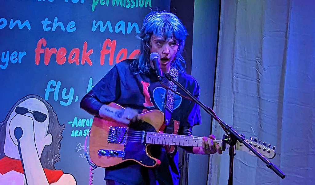A woman with blue hair holding a guitar and singing into a microphone.
