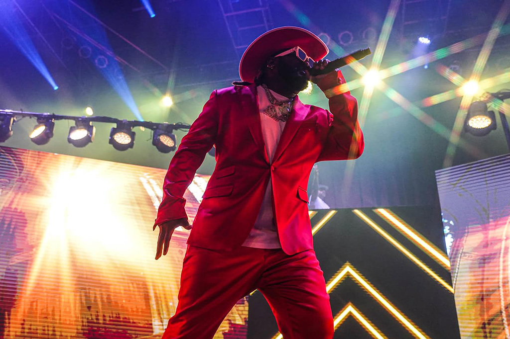 A rapper onstage with a red suit and red cowboy hat on holding a microphone near his mouth.