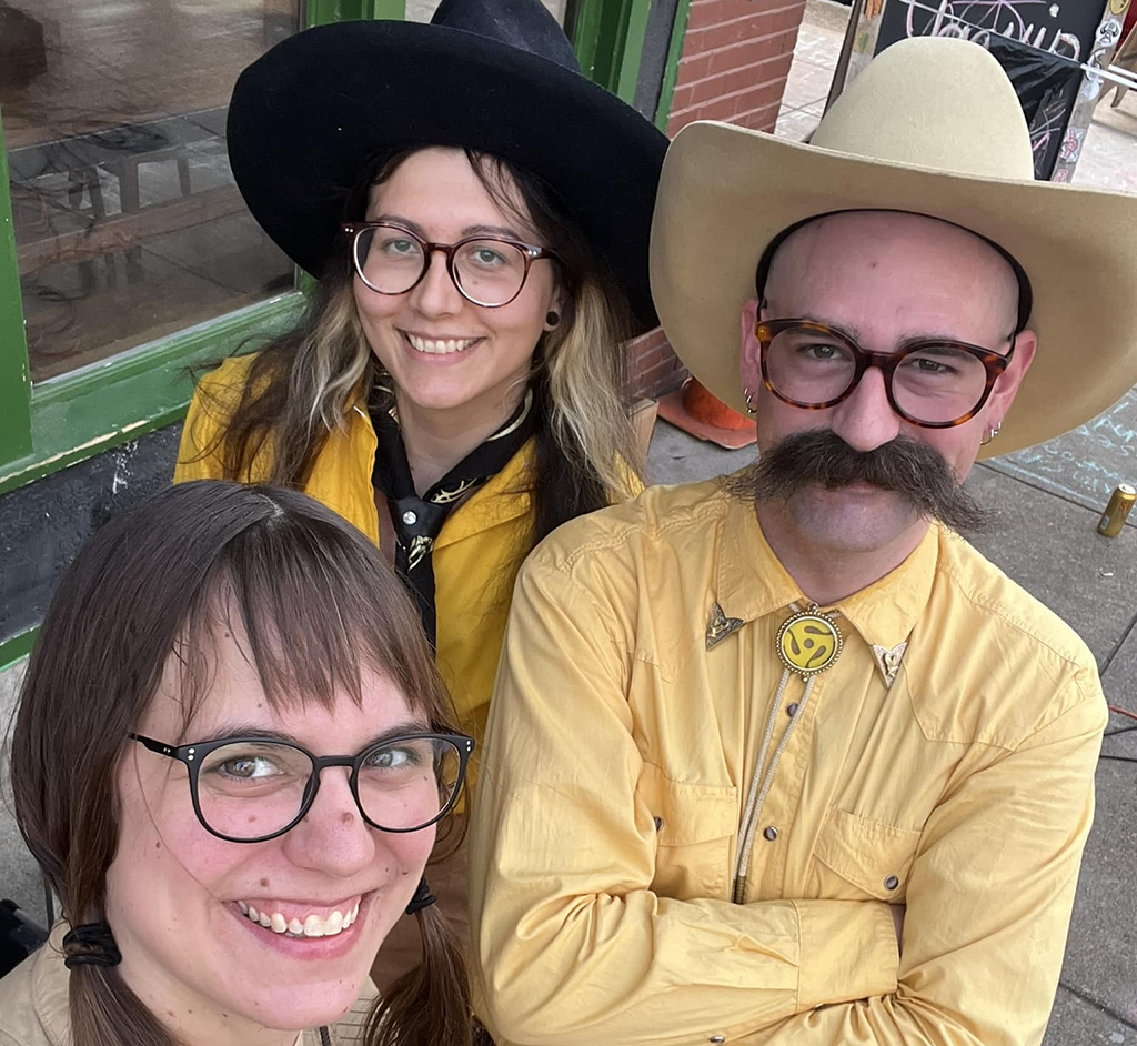 3 members of The Goldenrods posing for the camera on a side walk. Two members are wearing over-sized cowboy hats