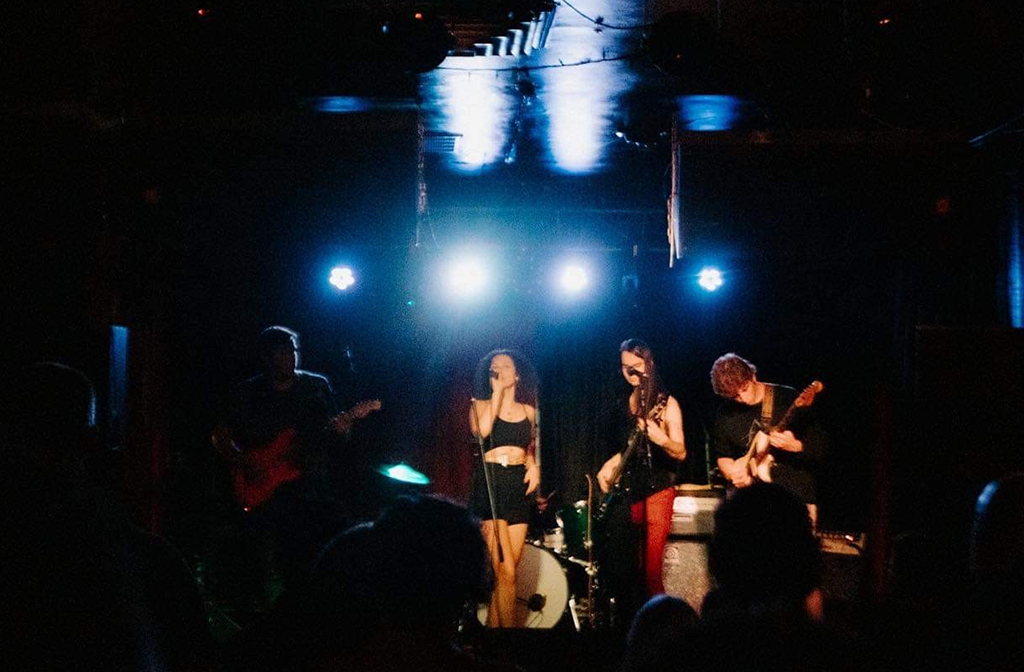 4 of the 5 members of The Hangovers performing on a stage that's dimly lit on the left. There is a male guitarist to the left, a female singer next to the right, a male bass player and a male guitarist. They are wearing mostly black clothing.