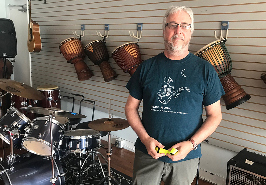 A man wearing a blue t-shirt that reads "Olde Music, Baroque and Renaissance Ensemble". He is standing in a room with many musical instruments.