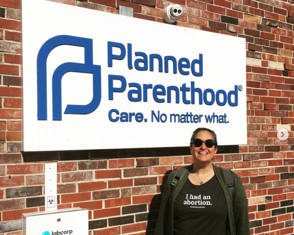 A white woman in a black t-shirt, zip up sweatshirt, and black sunglasses, stands in front of a brick wall with a planned parenthood sign. 