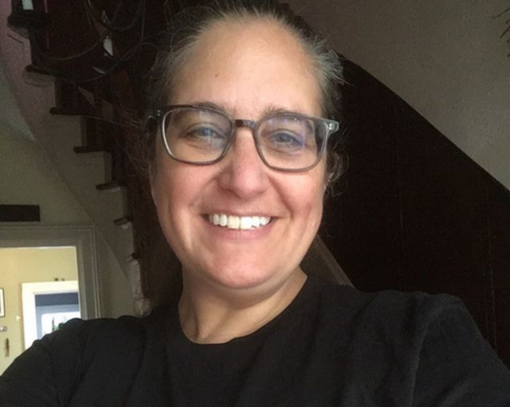 A picture of Julie Laut, a white woman with her hair pulled back, glasses, and a black t-shirt. She is smiling. 