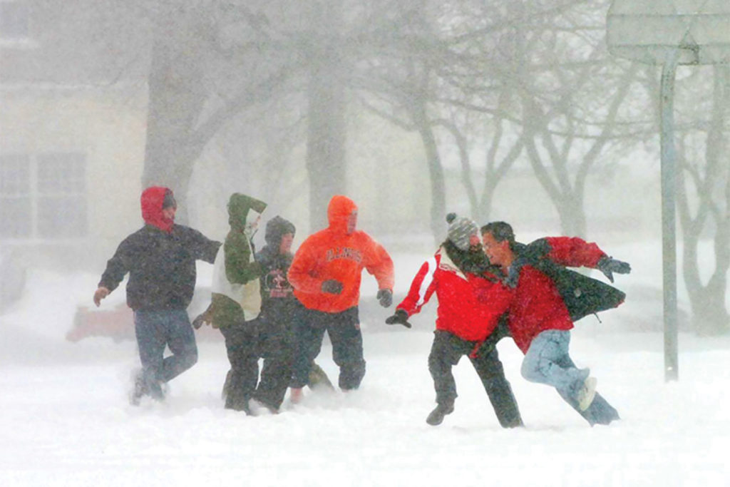 A group of students wearing winter coats, snow pants, hats and gloves run and play in deep snow while more snow comes down, blocking out most of the background behind them. 