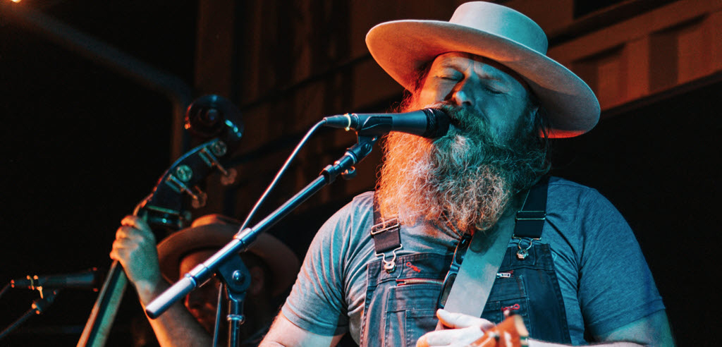 A man in a beard and a wide rim hat and overalls singing into a microphone and holding a guitar.