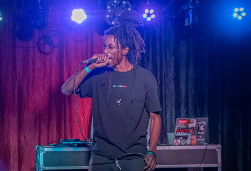 A rap artist onstage performing. He has long dreadlocks that are tied up in a bun and he's dressed in all-black.