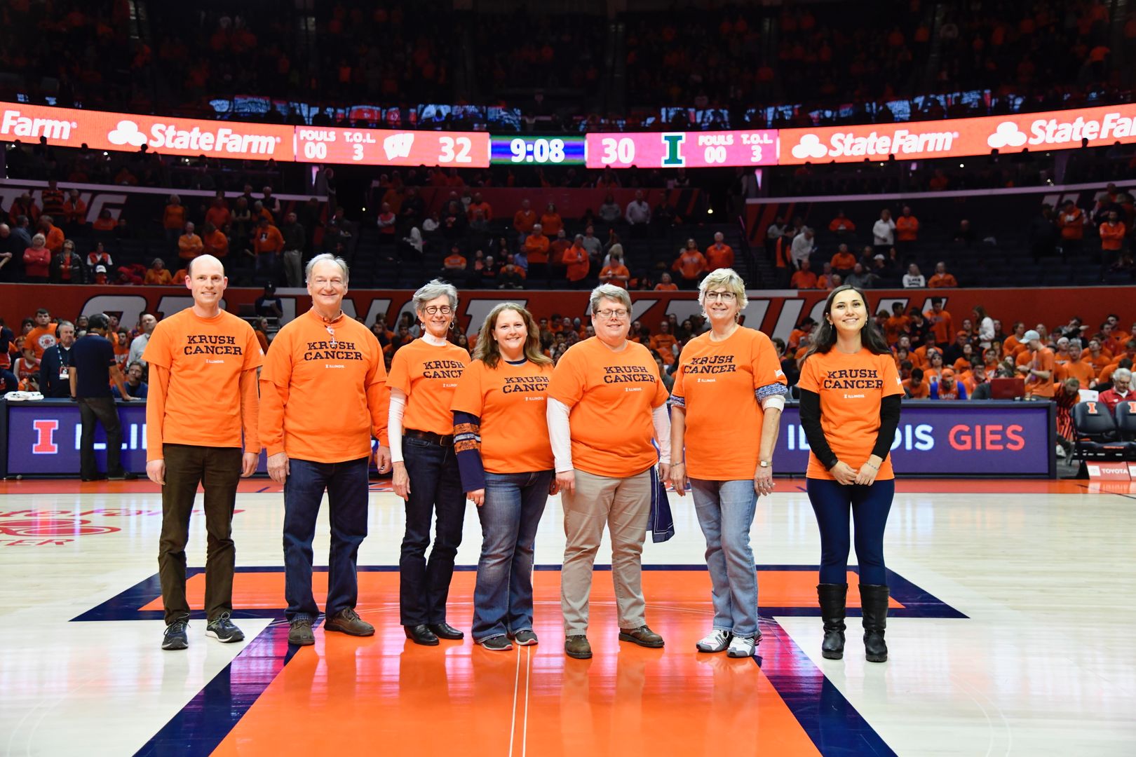 Seven people are standing in a line in the center of the basketball court at State Farm Center. They are wearing orange shirts that say Krush Cancer in blue.