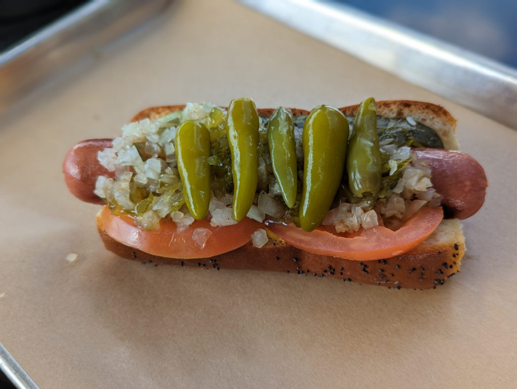 close-up of a loaded hot dog on grilled bun with poppyseeds, covered with tomatoes, minced onions, mustard, pickle and sport peppers. Photo by Tayler Neumann.