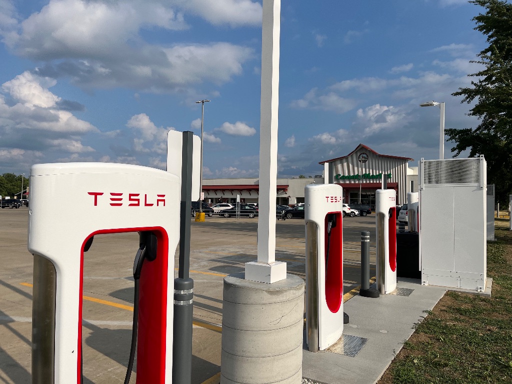 A row of white and red Tesla electric charging stations stand at the edge of a parking lot with the beige county market grocery in the background. It's a sunny day and the sky is blue with white fluffy clouds.
