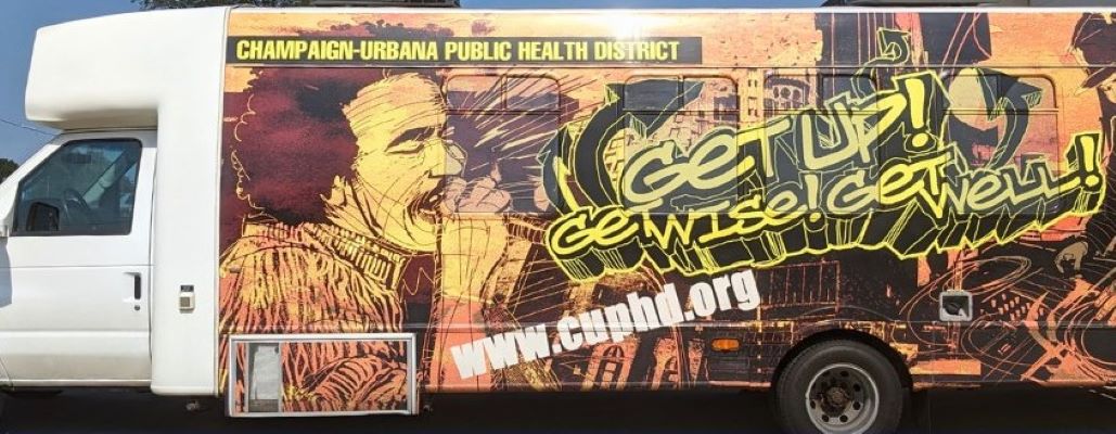 A large van with an illustration that covers the entire side of it. It depicts a DJ and a person singing, with a city skyline in the background. In yellow graffiti style letters it says Get Up, Get Wise, Get Well!