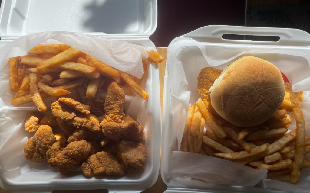 Da Shark: An overhead picture of two to-go containers. Inside the container at the top is a sandwich on a bun with a side of french fries. The bottom container holds fried fish nuggets and a side of fries. Photo by Remington Rock.
