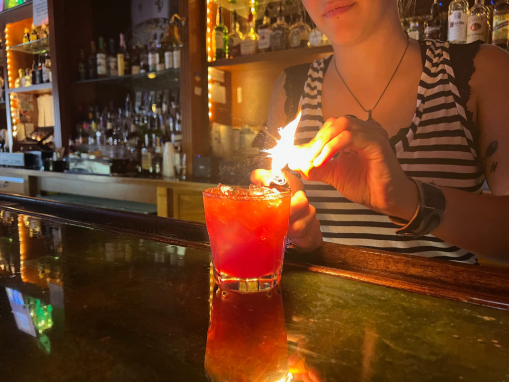 Bartender at Bentley's Pub Katie Carrillo flames an orange peel over an old fashioned cocktail. The flake is bright and over-exposed, and the drink is bright red. Photo by Alyssa Buckley.