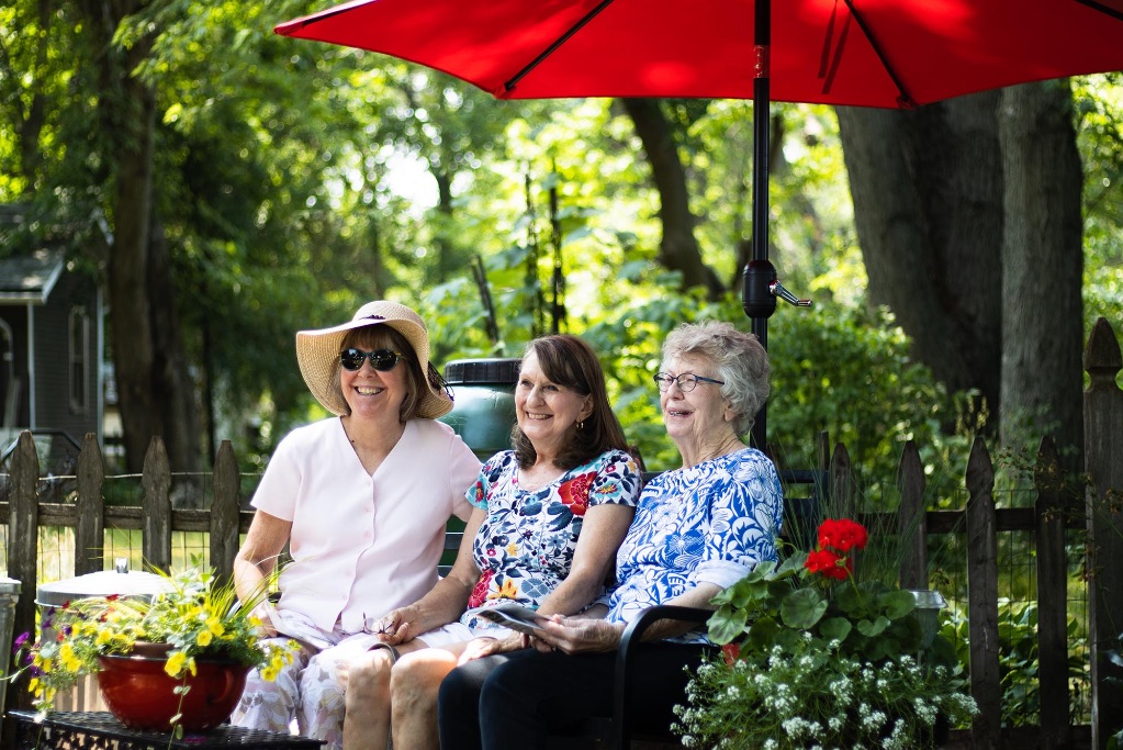 3 white women sit together on a bench under a red umbrella on a sunny day. They are smiling. The woman on the left is white and wears a large sun hat and sunglasses and a white shirt. The woman in the middle is white with brown shoulder length hair and wears a printed shirt with blue, red, and white colors. The woman on the right is also white and has gray and brown hair and a blue and white shirt.