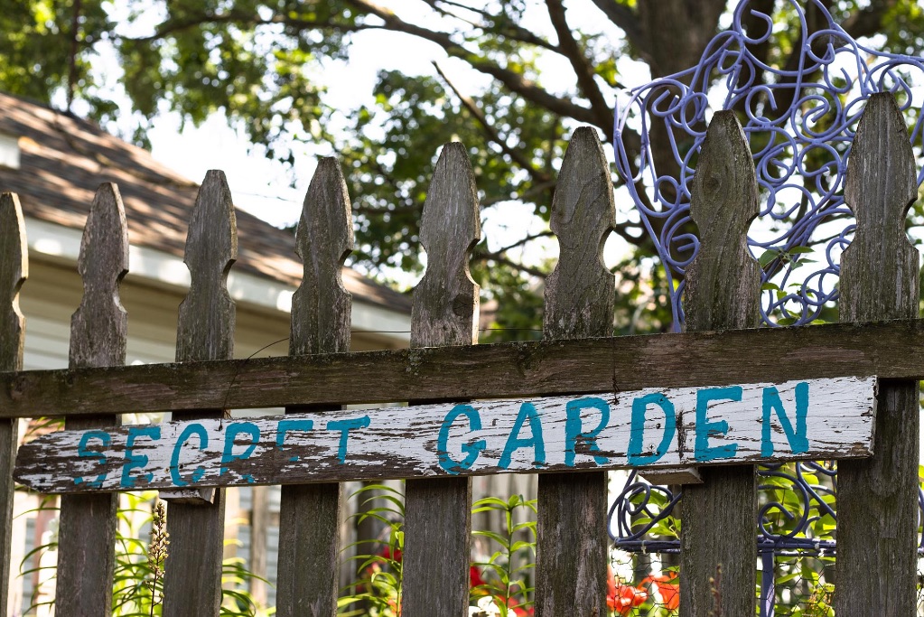 An image of a brown wooden fence with a well worn sign with chipped white paint and blue hand painted letters that spell out "secret garden"