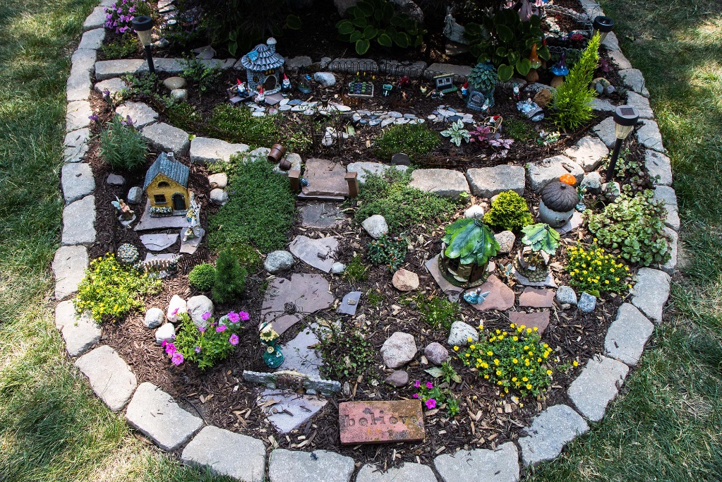 An image of a fairy garden. The garden is surrounded by gray bricks, with tiny houses, flowers, and gnomes. There is a stone in the center that says believe. 