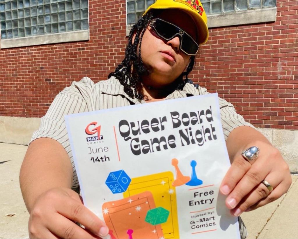 A person with shoulder length black hair, sunglasses, and an orange hat holds a piece of paper up to the screen that says Queer Board Game Night.