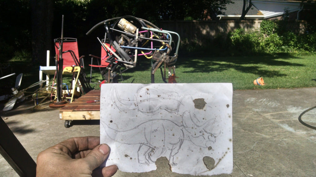Andy White holds a piece of paper in the foreground of a sketch of his dinosaur sculpture "Beauty". In the background, the sculpture is in its early stages with just a basic shape taking form. The sculpture is outside. 
