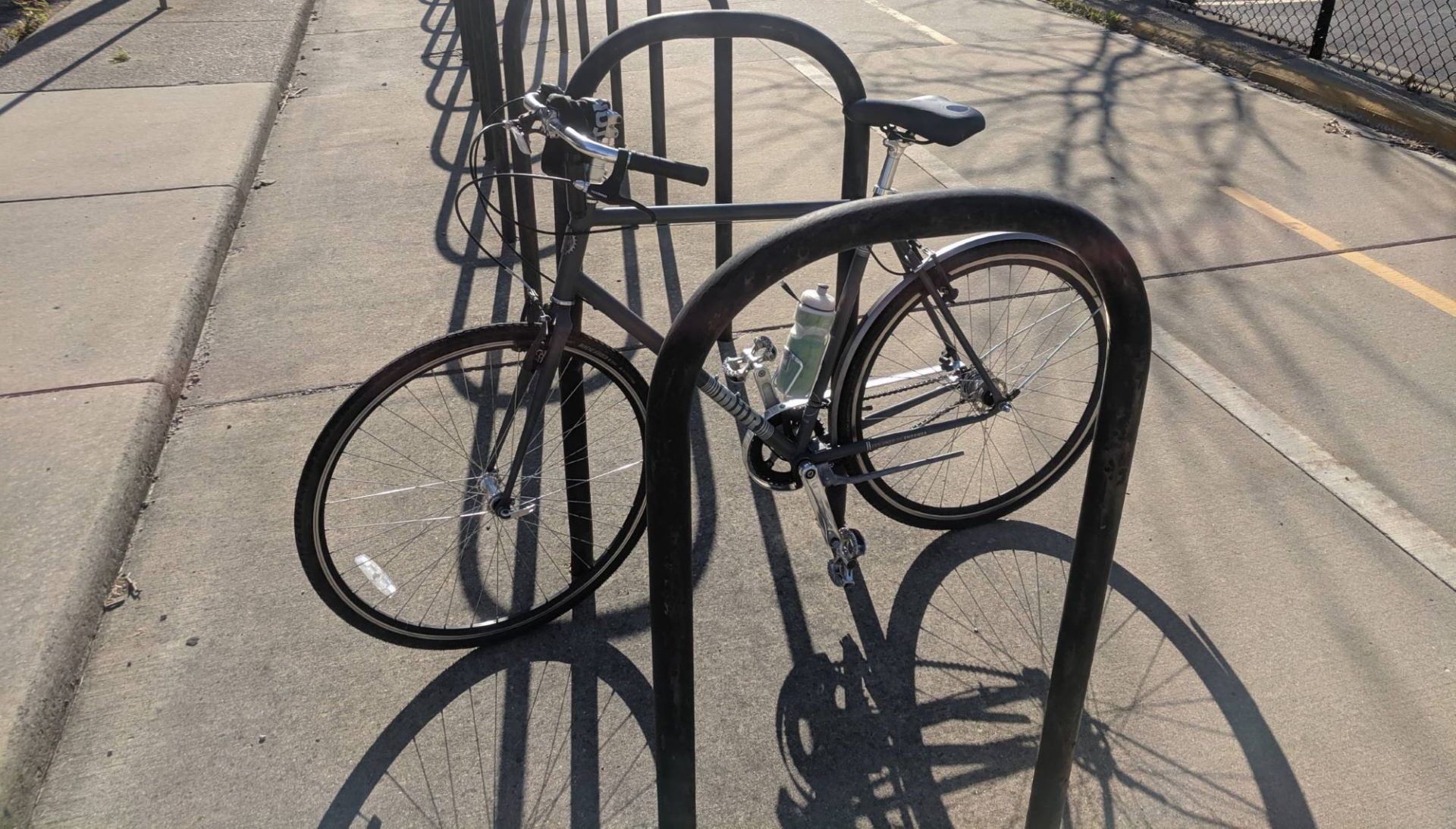 A bicycle is attached to a bike rack with several U shapes made of black metal. The sun is shining and creating a shadow of the bike on the concrete.