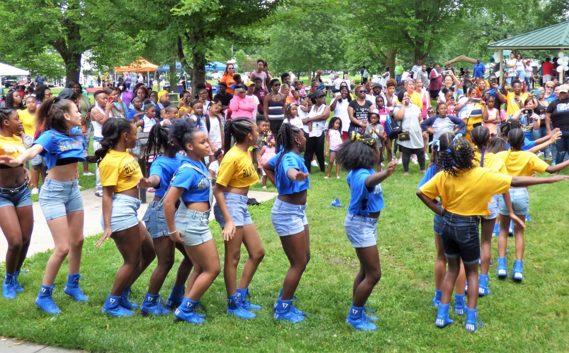 Several young Black girls in blue and yellow t-shirts and matching blue shoes are in a line doing a dance. There is a crowd of people looking on.