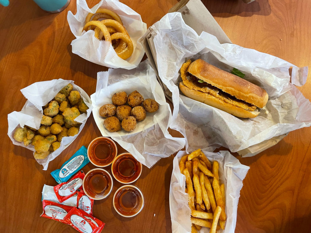 Big JJs: an overhead shot of baskets of food. Includes a basket of hushpuppies, fried okra, onion rings, and a sandwich. Next to the basket are packets of sauce. Photo by Remington Rock.