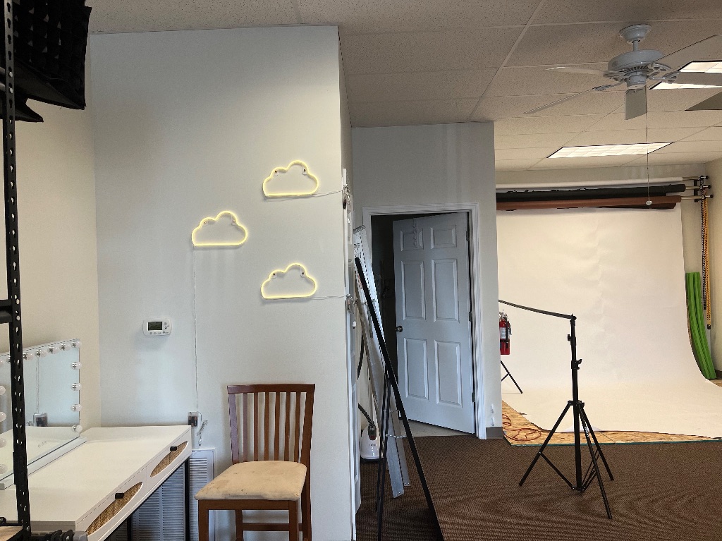 The wall of a studio, with neon clouds, a white backdrop, and a wooden chair in front of a table sit on the left side of the picture.