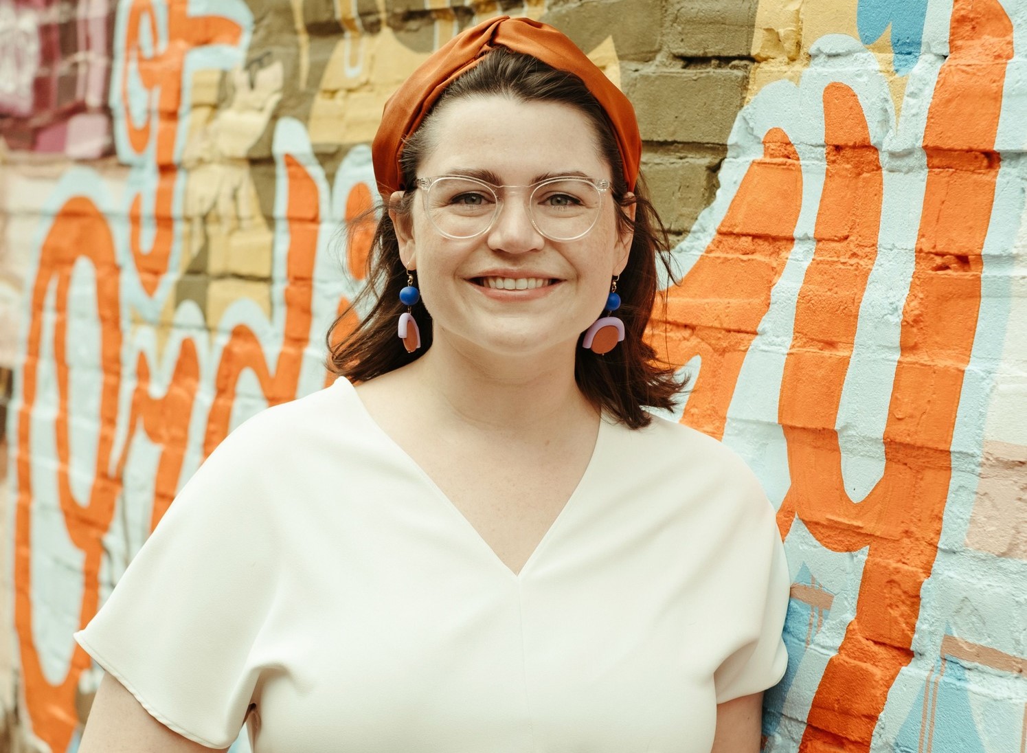 A white woman with short brown hair and an orange headband is leaning against a brick wall. The wall is painted with a colorful mural.