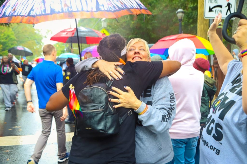 A white woman with blonde hair is hugging Black person with a black shirt and black backpack. They appear to be at a parade. Several people around them are holding umbrellas.