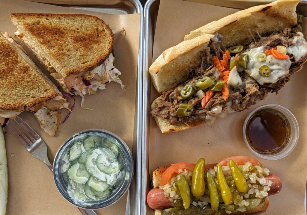 Food items sit on two metal trays covered in brown paper. On the left tray is a toasted sandwich cut in half with meat and sauce spilling out. The tray also contains a fork and an unopened plastic container with a cucumber salad. At the top of the right tray is an open Italian beef sandwich with papers, cheese, and beef visible as well as a cup of dipping au jus sauce. At the bottom is a Chicago-style hot dog covered in pepper and onions. Photo by Tayler Neumann.