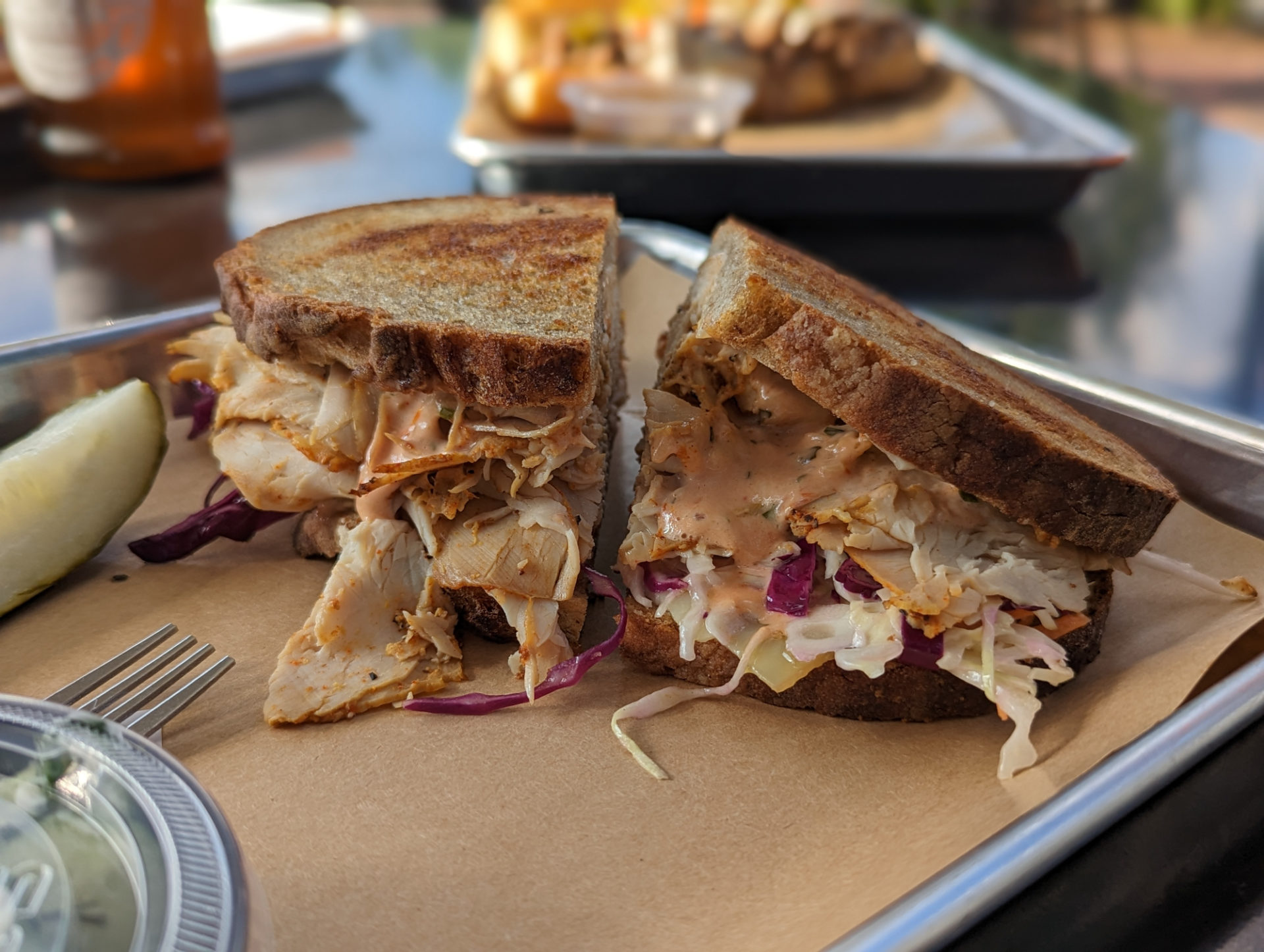A side view of a sandwich on a tray. Toasted bread with turkey, sauce, and coleslaw spilling out. The tray also has a fork, a deli container, and a pickle. In the background, out of focus, there are other trays of food at Martinelli's Market. Photo by Tayler Neumann.