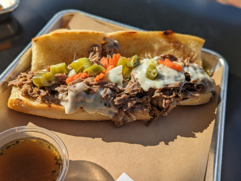 A close-up of an Italian beef sandwich on a baguette. Peppers, cheese, and beef are all visible. On the tray is a small plastic cup of au jus dipping sauce at Martinelli's Market in Champaign, Illinois. Photo by Tayler Neumann.
