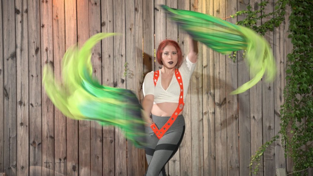a burlesque dancer in grey pants and a white crop top with orange suspenders waves large green fabric in the air. 