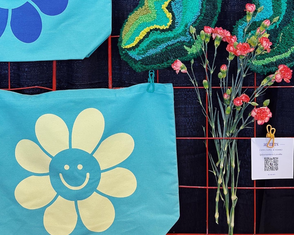 Two blue bags with a yellow and a blue daisy flower on it with smiling faces hang on a red wire rack. There are pink carnations in a vase next to them.