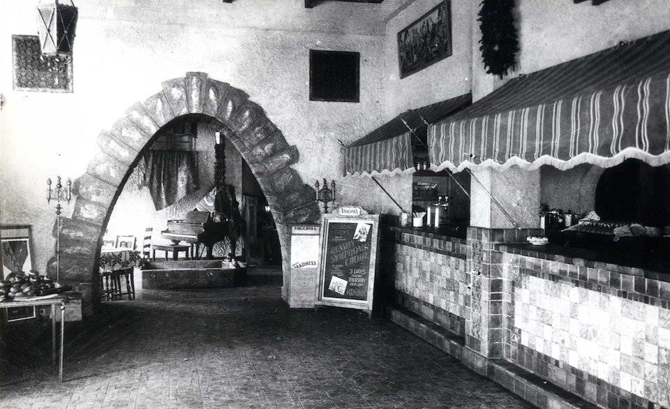 A black and white photo of the interior of a restaurant. There is a counter to the right, with awnings over it. There is an archway lined with bricks that leads into a room with a dining area.