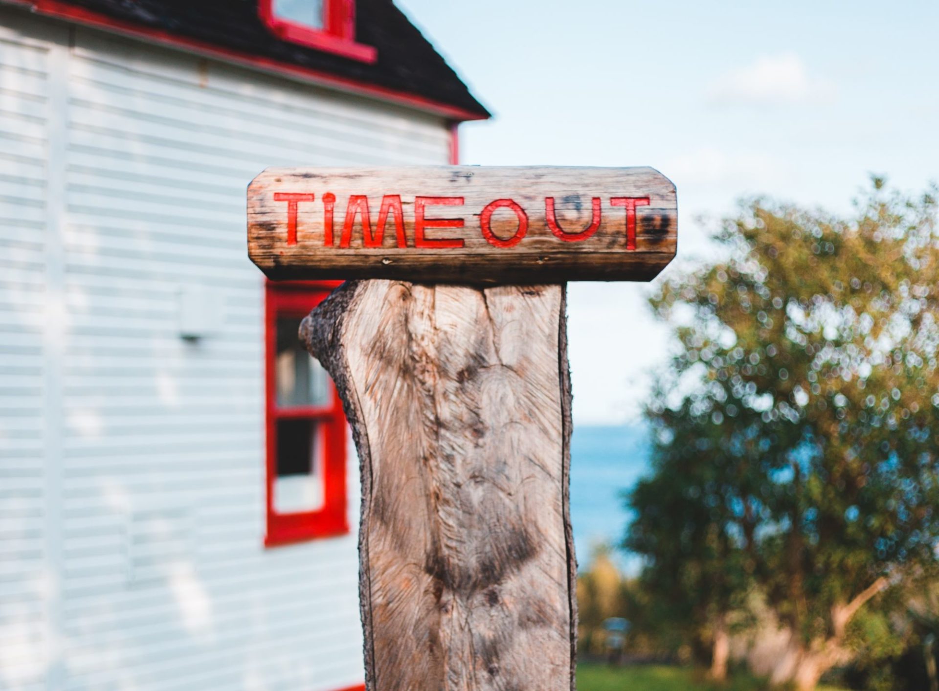 A log, cut in half standing vertically with a wood sign that has the words Time Out carved in it, painted red. It sits in front of a house with white siding and red window frames.