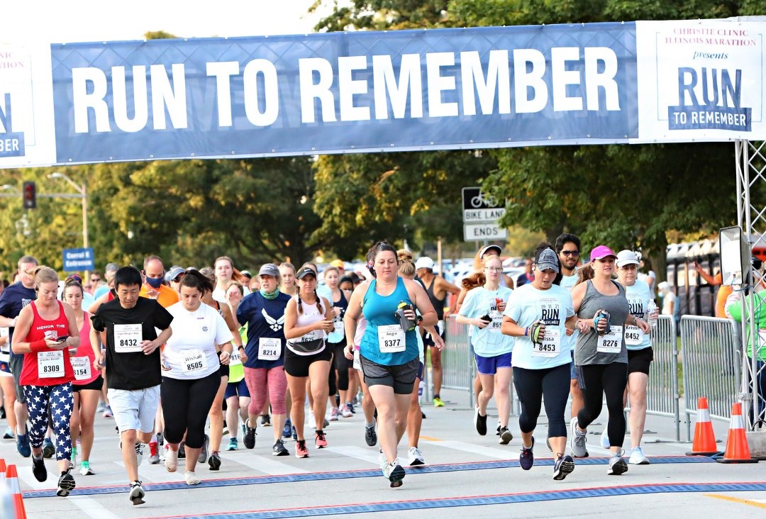 A group of people at the start of a running race. The are running under a large blue and white banner that says Run to Remember.