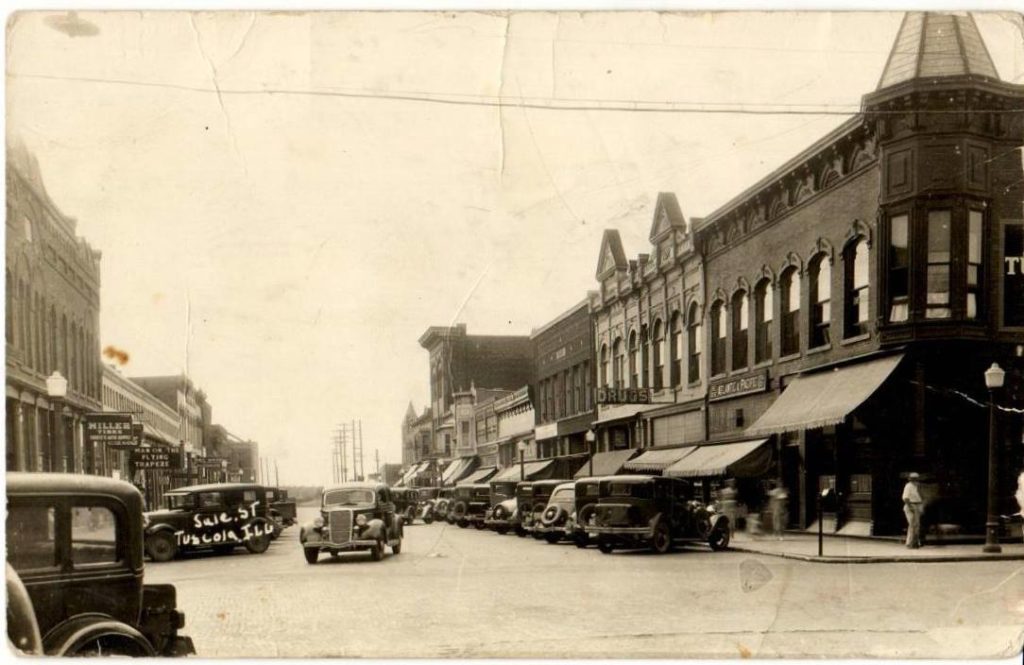A sepia toned photo from the 1930s depicting a downtown block lined with buildings and cars. 