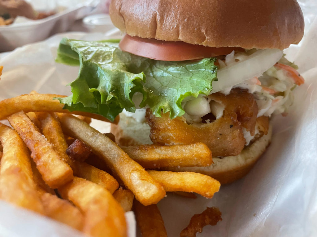 A closeup of a sandwich and french fries. The sandwich is layered with a fried filet, coleslaw, onions, lettuce, and tomato. Photo by Remington Rock.
