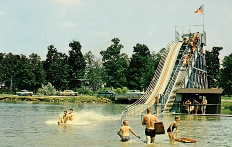 An old photograph of a tall slide that extends into a lake. There are people in the water surrounding it, as well as standing in line on the stairs.