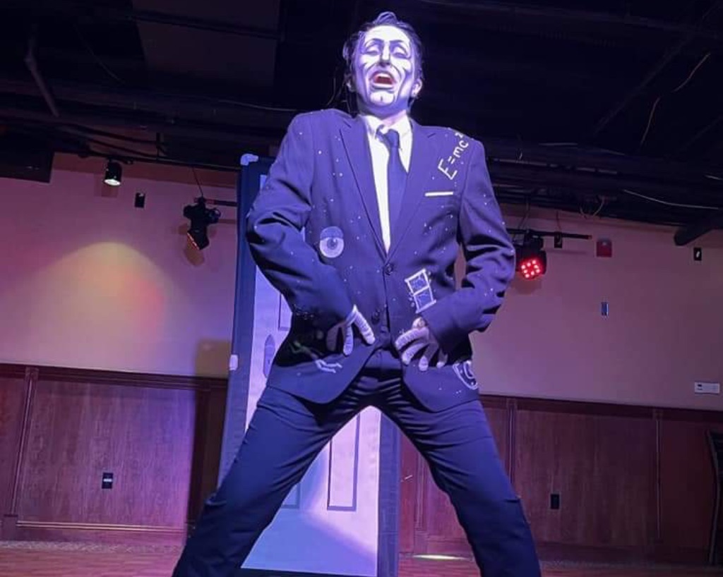 Performer Spank Knightly stands on stage. He wears a navy suit and tie, The jacket has images of an eyeball, window, and E= MC2. He has his hands on his waist.His face is painted white with black lines to give the illusion of a mask.