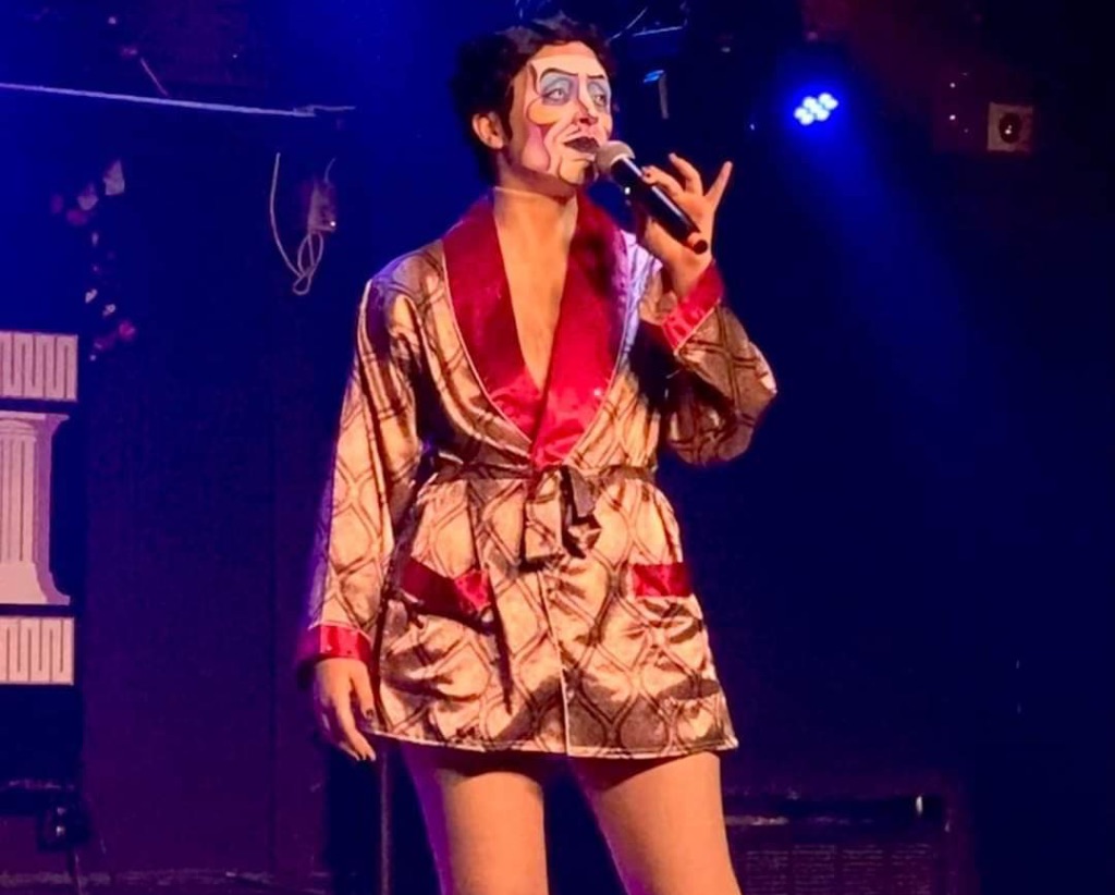 Performer Spank Knightly stands on stage. He wears a tan and red silk robe. He is holding a microphone.His face is painted white with black lines to give the illusion of a mask.
