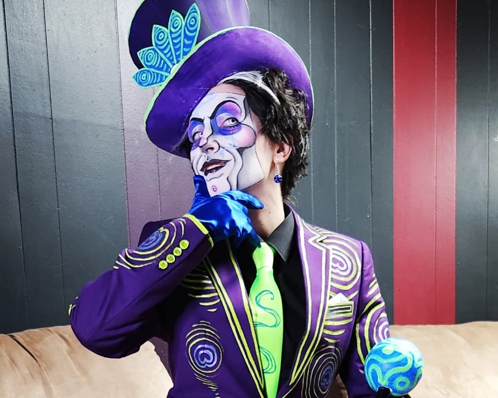 Performer Spank Knightly sits on a white sofa. He wears a purple painted suit jacket, a neon green tie, blue gloves, and a purple top hat. His face is painted white with black and purple lines to give the illusion of a mask.