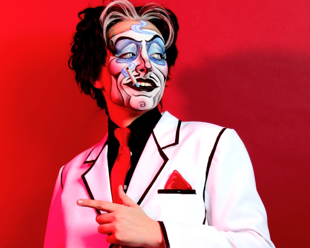 Performer Spank Knightly stands in front of a red backdrop. He wears a white suit with black trim and a red pocket square and tie. His face is painted white with black lines and blue eyeshadow to give the illusion of a mask. He has black and white hair. He has a cigarette in his mouth.