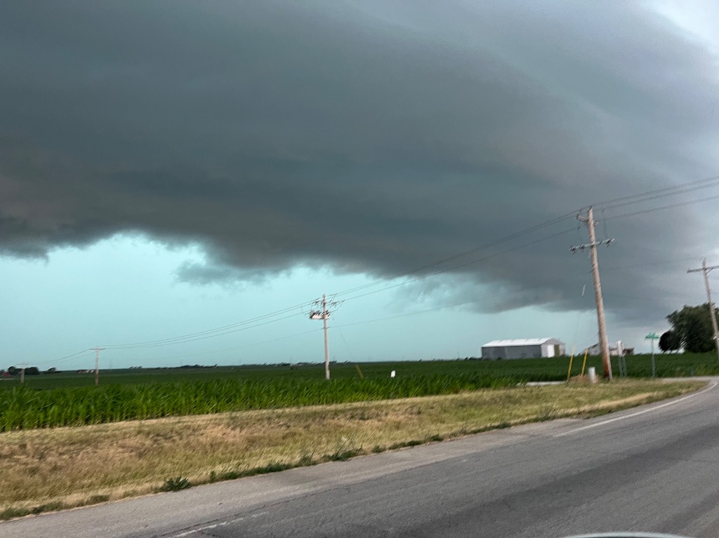 A huge dark gray storm front moves through the sky. The wall of clouds moves through the sky over a field of green plants and a bit of road in the lower right corner.