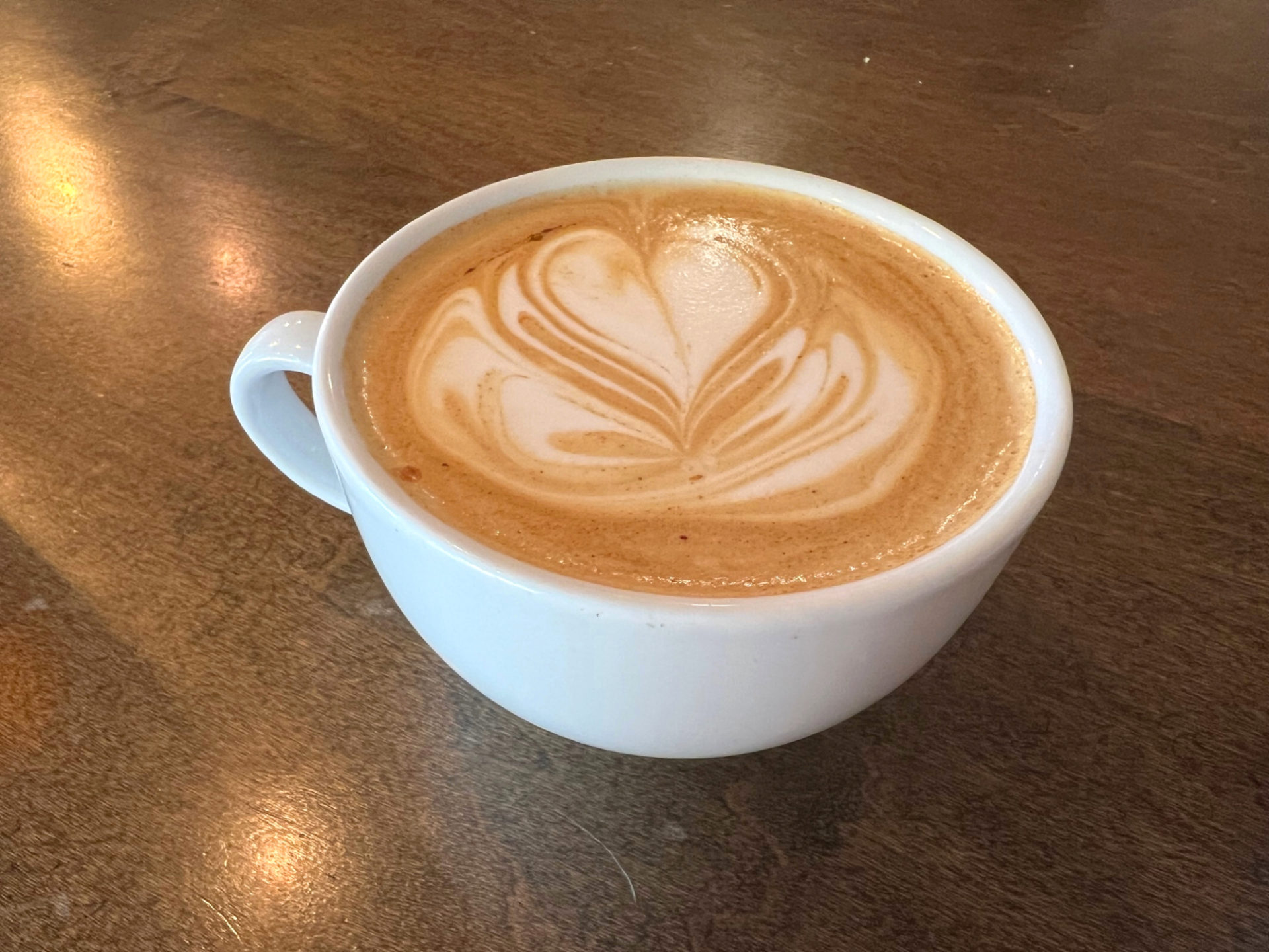 Sweet heat latte from Avionics for five things to eat or drink in Champaign-Urbana for June 2023. A light brown beverage in a white mug on a dark brown wood table. There is white latte art designed in the drink.