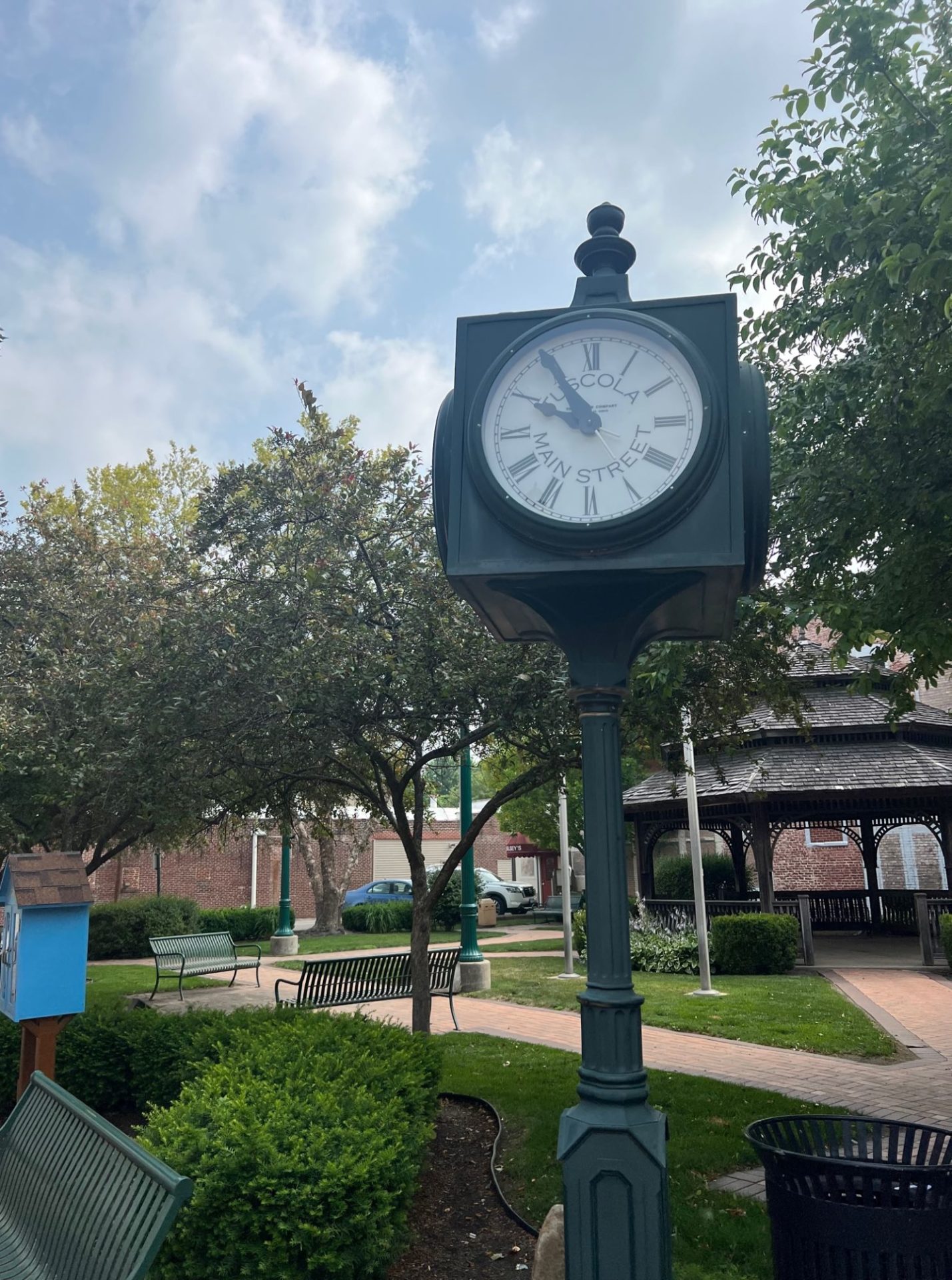 An old-fashioned black post with a white clock face stands at the corner of a plaza with a gazebo and brick sidewalks running through it.