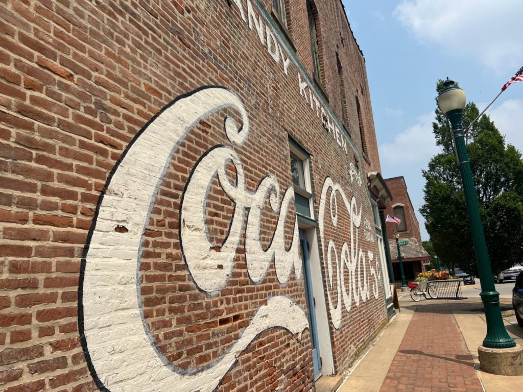 A brick facade of a building that is painted with a large white Coca-Cola logo. A red brick sidewalk runs alongside it, and there is a green lampost across from the mural.