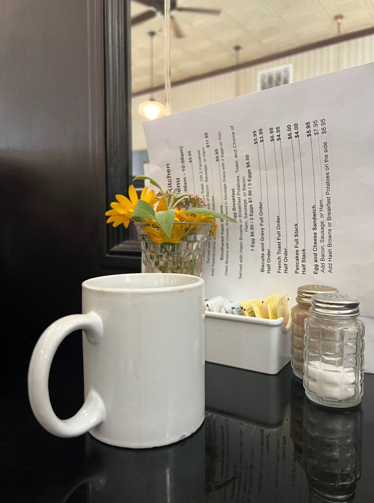 A white coffee mug sits on a dark brown table, alongside a vase of yellow flowers, a container of sugar packets, salt and pepper shakers, and white paper menus.