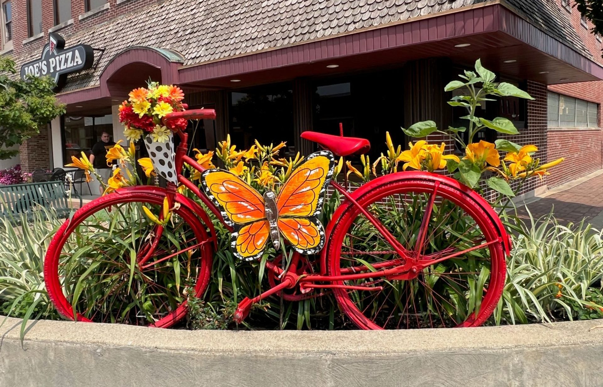 A cement planter is filled with colorful flowers, and a red bicycle decoration with an orange and yellow decorative butterfly.