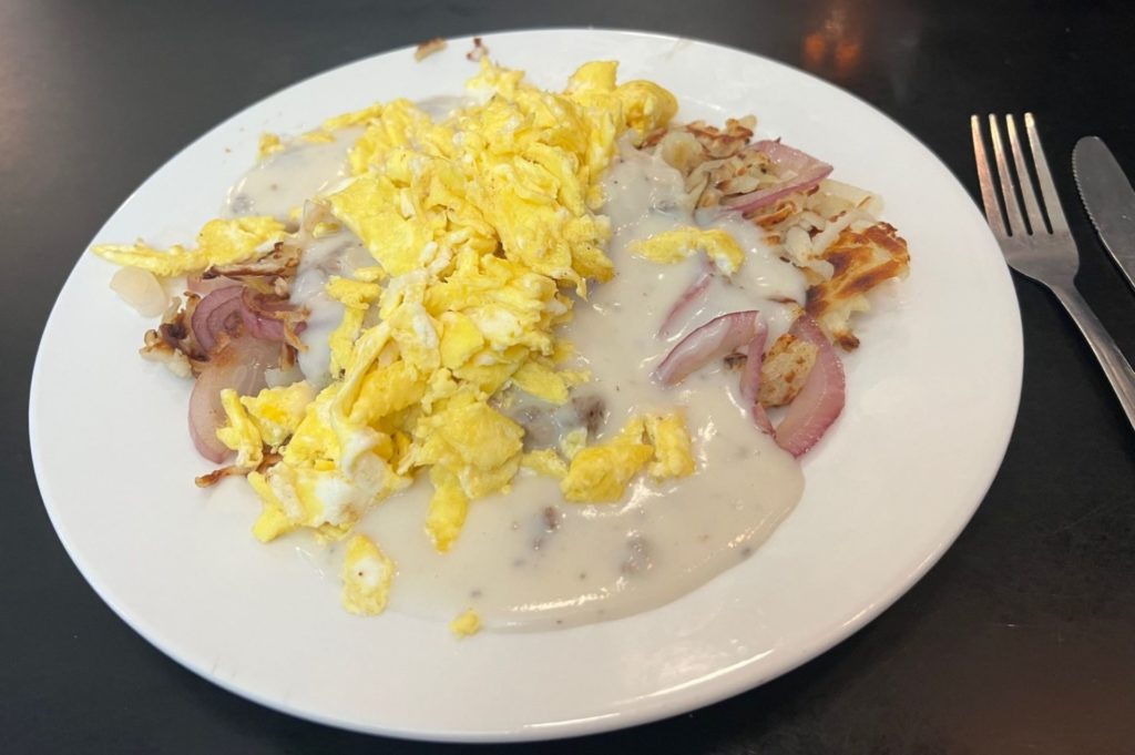 A round white plate that has shredded potatoes, onions, white gravy, and scrambled eggs piled on it. It sits on dark brown table, and there is a fork and knife next to it.